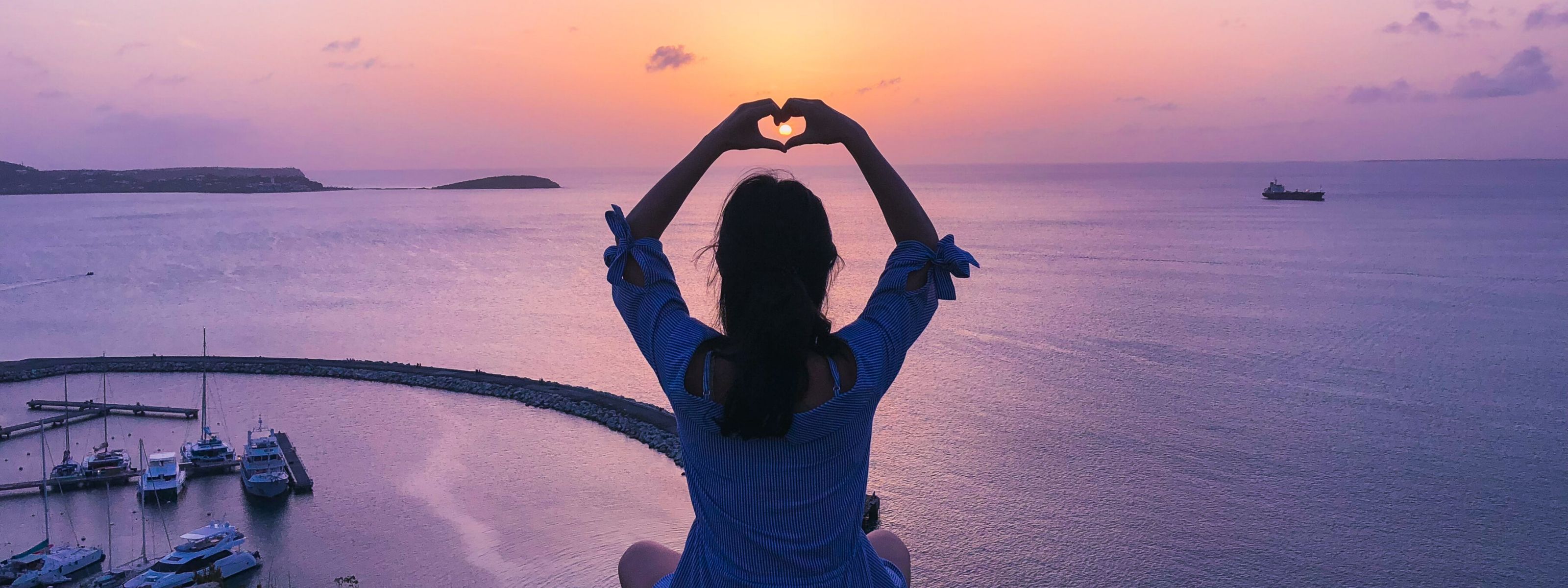 Kobieta układa dłonie w kształt serca na tle zachodu słońca nad morzem.