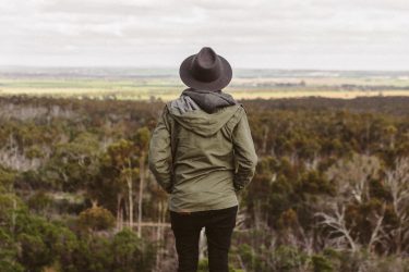 Kobieta w kapeluszu wpatrzona w horyzont. U jej stóp las.
