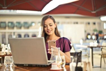 Kobieta siedzi przy stoliku w kawiarni, korzysta z laptopa i smartfona