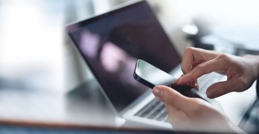 Zbliżenie na dłonie korzystające ze smartfona przy otwartym laptopie – mobilna karta SIM do biura i pracy w terenie.