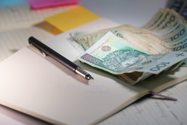 Długopis leżący na zeszycie w towarzystwie banknotów polskich - ilustracja obliczania należnego wynagrodzenia uczniów