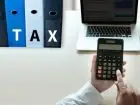 Segregatory z napisem "tax", laptop na blacie i kalkulator w dłoniach