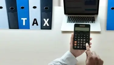 Segregatory z napisem "tax", laptop na blacie i kalkulator w dłoniach