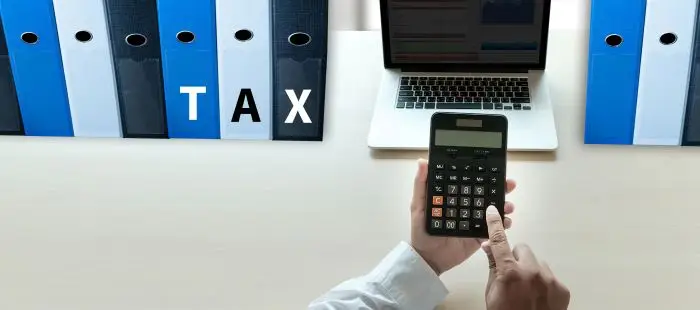 Segregatory z napisem "tax", laptop na blacie i kalkulator w dłoniach