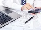 Koszty uzyskania przychodów - pracownik liczy na kalkulatorze siedzi przy biurku na którym stoi laptop oraz leżą rozliczenia PIT