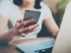 Telefon służbowy na urlopie - kobieta trzymająca telefon w dłonie siedząc przy biurku przed laptopem, patrzy na ekran