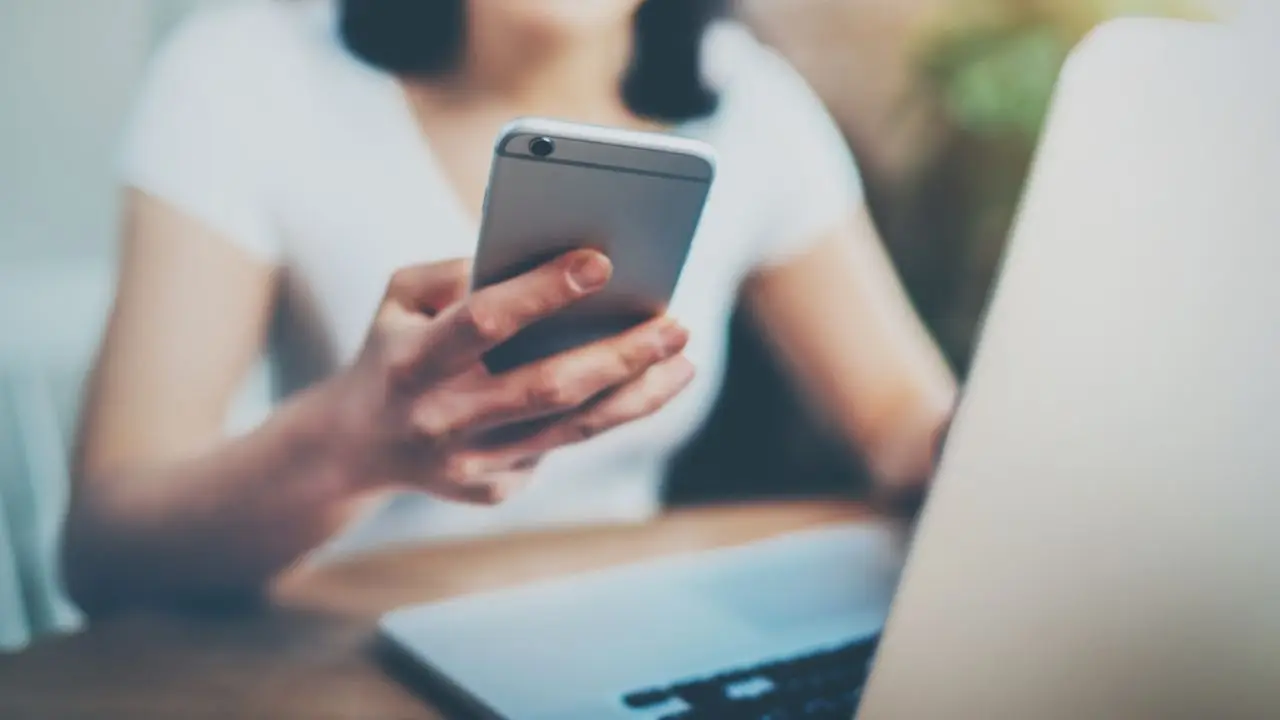 Telefon służbowy na urlopie - kobieta trzymająca telefon w dłonie siedząc przy biurku przed laptopem, patrzy na ekran