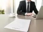 Mężczyzna siedzący przy biurku, pracujący z laptopem. Na biurku leży umowa