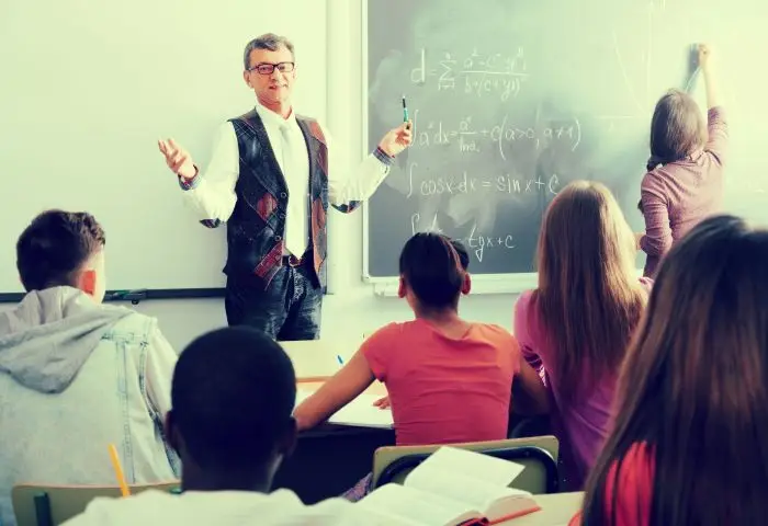 Nauczyciel stojący przy tablicy w sali lekcyjnej, patrzą na niego dzieci w ławkach