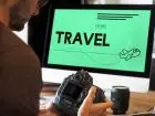 Meżczyzna trzymający aparat, siedzący przed kompyetrem, na ekranie napis "travel"