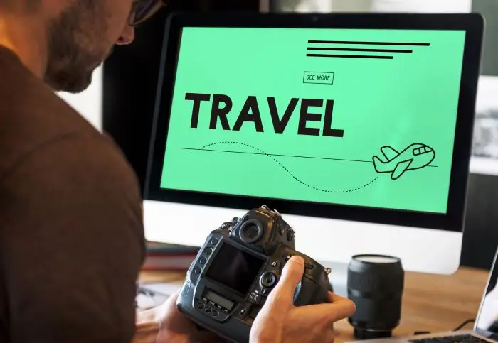 Meżczyzna trzymający aparat, siedzący przed kompyetrem, na ekranie napis "travel"