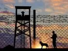 Straż graniczna - widok ogrodzenia z drutem kolczastym, wieżyczki strażniczej oraz strażnika granicznego z psem na tle zachodzącego słońca