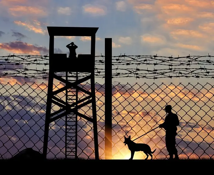 Straż graniczna - widok ogrodzenia z drutem kolczastym, wieżyczki strażniczej oraz strażnika granicznego z psem na tle zachodzącego słońca