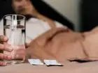 Osoba leżąca w łóżku, zbliżenie na szklankę wody i tabletki