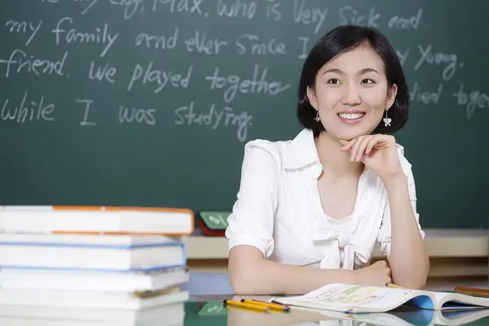 Nauczyciel dyplomowany - uśmiechnięta młoda nauczycielka siedzi przy biurku na tle tablicy szkolnej