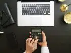 Zbliżenie na dłonie podczas dokonywania obliczeń na kalkulatorze, w tle laptop