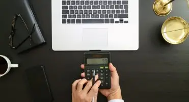 Zbliżenie na dłonie podczas dokonywania obliczeń na kalkulatorze, w tle laptop