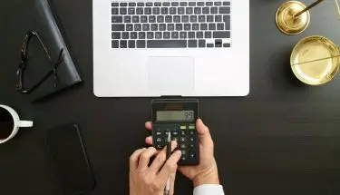 Zbliżenie na dłonie podczas dokonywania obliczeń na kalkulatorze, w tle laptop