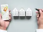 Inwestowanie w nieruchomości bez pieniędzy - rozłożone dłonie nad trzema figurkami domów. W jednej dłoni mapa, w drugiej długopis