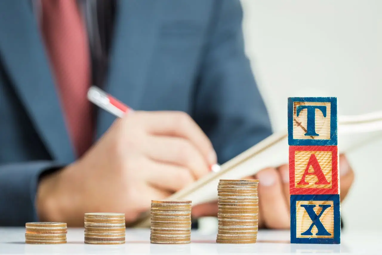 Kto nie może być na ryczałcie 2024 - mężczyzna dokonujacy obliczeń w notatniku, obok niego stosy monet. Napis z klocków "tax"