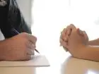 Testy behawioralne - dłonie dwóch osób siedzących na przeciwko siebie przy stole. Jedna osoba notuje