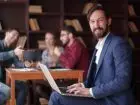 Co wpływa na wizerunek pracodawcy - mężczyzna siedzący z laptopem na kolanach, uśmiechnięty, elegancki. W tle trzy osoby omawiające dokumenty przy stole