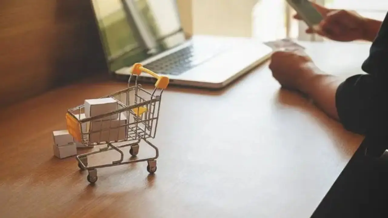 Duże zaległości e-handlu - miniaturowy wózek zakupowy na biurku, w tle osoba dokonująca płatności za pośrednictwem laptopa